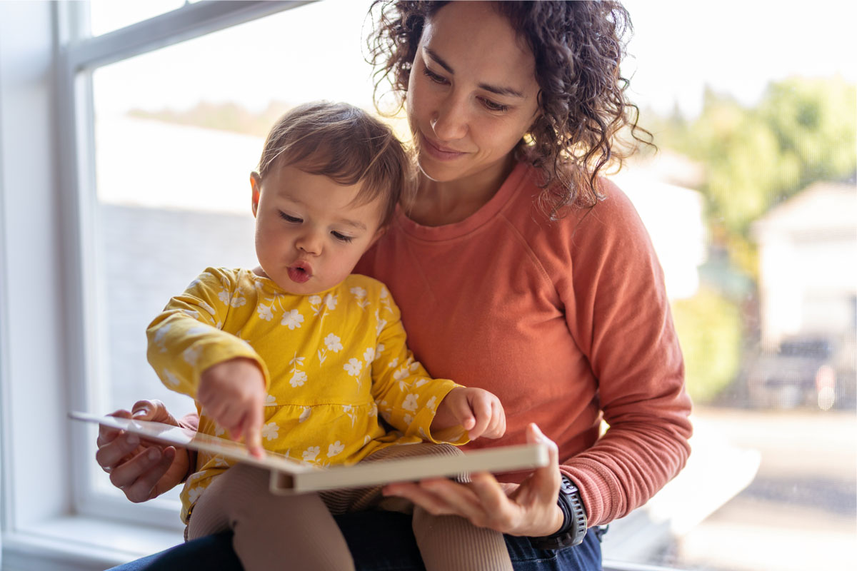 Mother and toddler sitting in front of a window, reading a book.